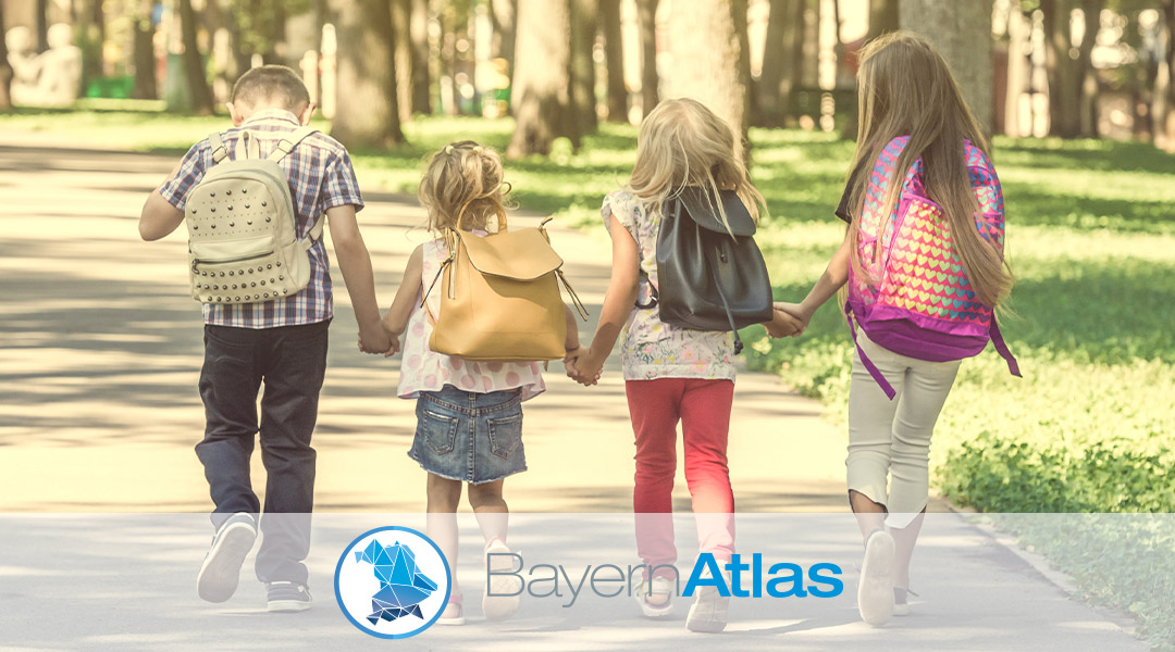 BayernAtlas-Thema Schulweg Vier Kinder mit Rucksäcken gehen Hand in Hand auf einer Straße.