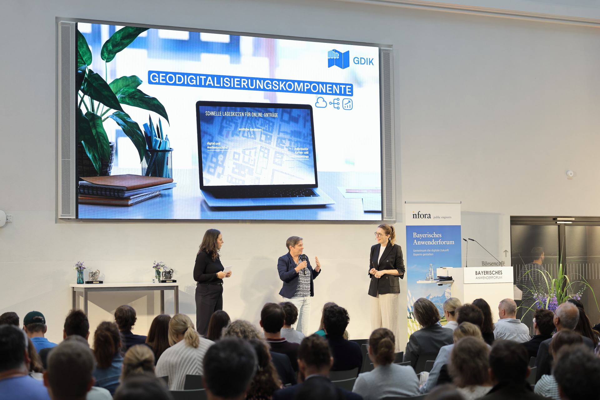 Digitale Verwaltung in Bayern "Bavarian Best Practice Award" 3 Frauen auf der Bühnen,  im Hintergrund an der Wand ein großer Bildschirm, vor der Bühne das Publikum